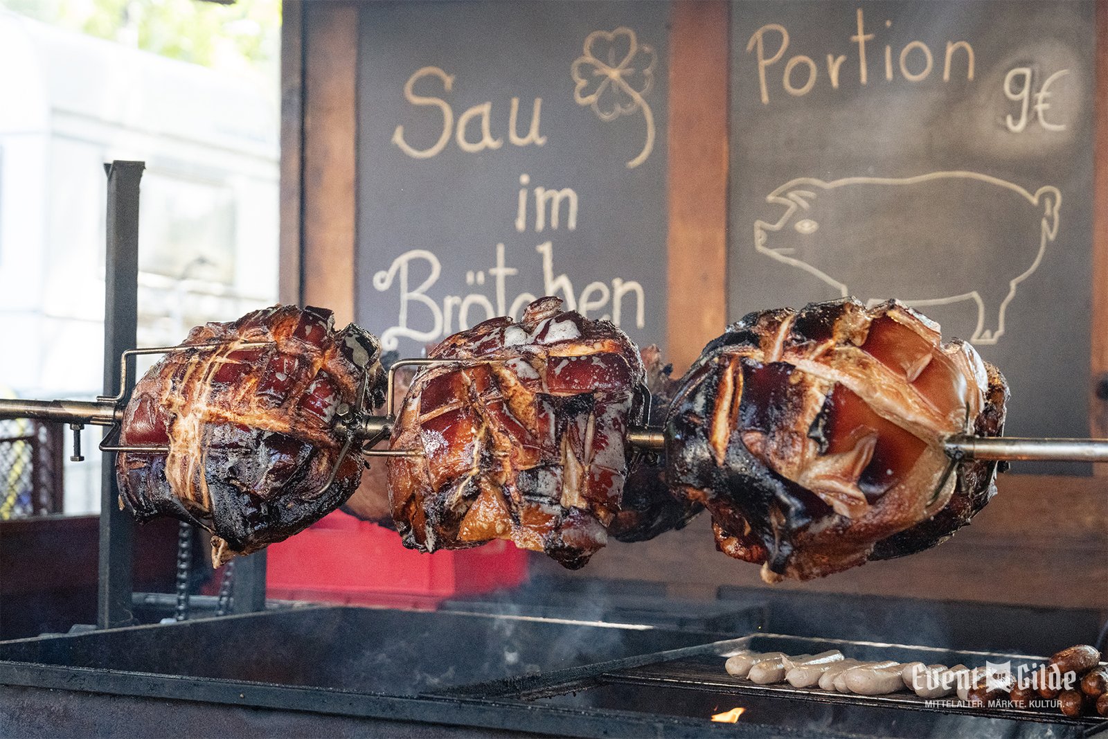 Schweinebraten am Spieß über der Glut, dahinter die Tafel mit der Aufschrift Sau im Brötchen für 9 Euro