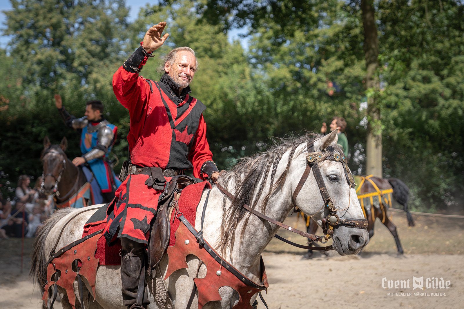 Ritter in rotem Wappenrock grüßt vom Schimmel aus das Publikum am Turnierplatz in Telgte