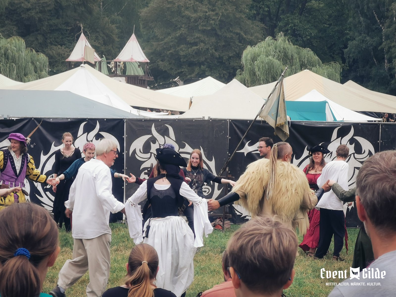 Gäste und Spielleute tanzen beim Tanzworkshop im Kreis auf der Wiese des MPS Bückeburg 2026