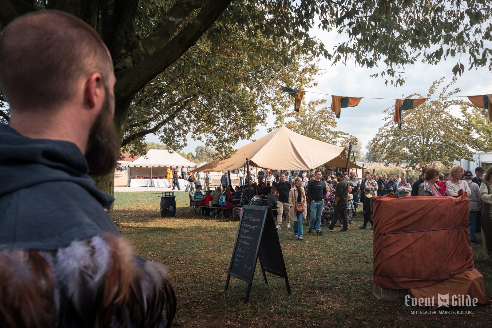 Gäste zwischen Zelten, Wimpelketten und einer Kreidetafel mit Speisen beim Wikingerfest an der Xantener Südsee, im Vordergrund ein gewandeter Besucher
