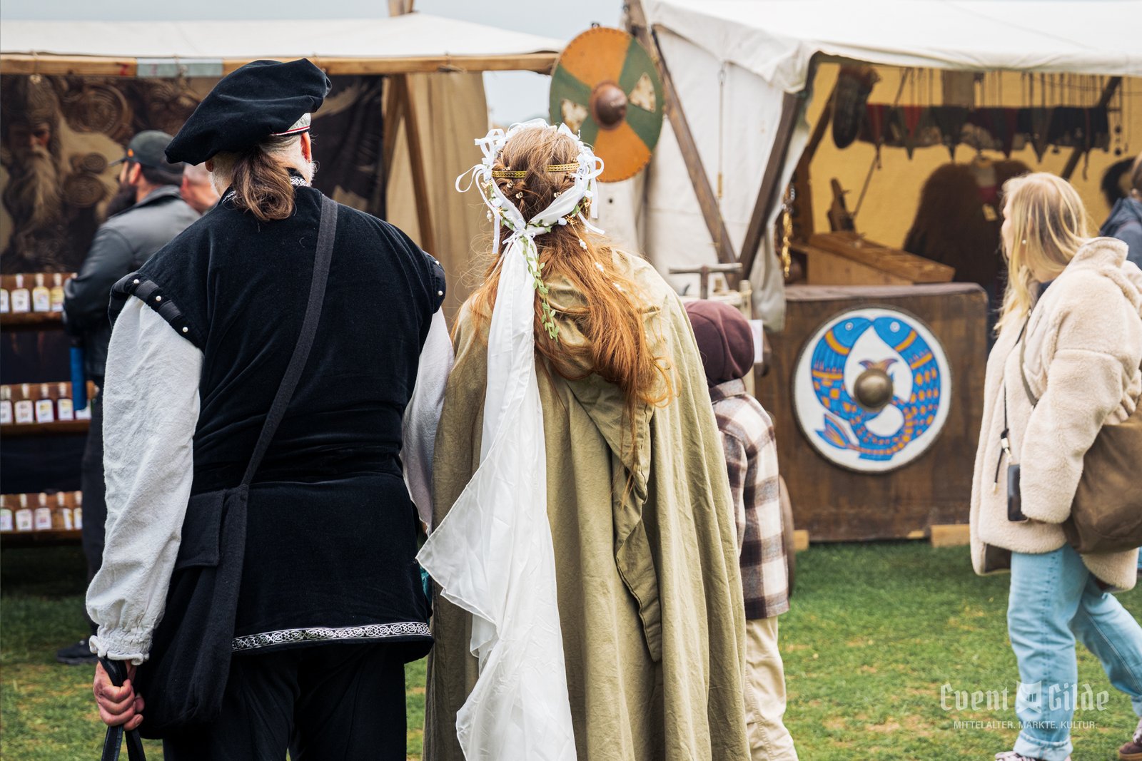 Gewandetes Paar mit Barett und Umhang schlendert an Ständen mit bemalten Rundschilden beim Wikingerfest in Xanten vorbei